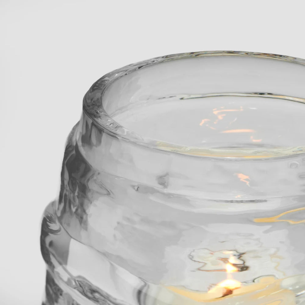 Close-up detail of a clear textured glass jar with a warm yellow light glowing inside, set against a plain light gray background.