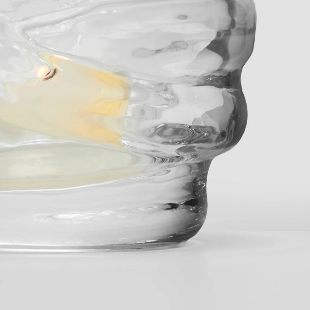 A close-up of a sculptural, organic-shaped clear glass vessel or candle holder with a small warm yellow glow visible inside, set against a clean light gray background.