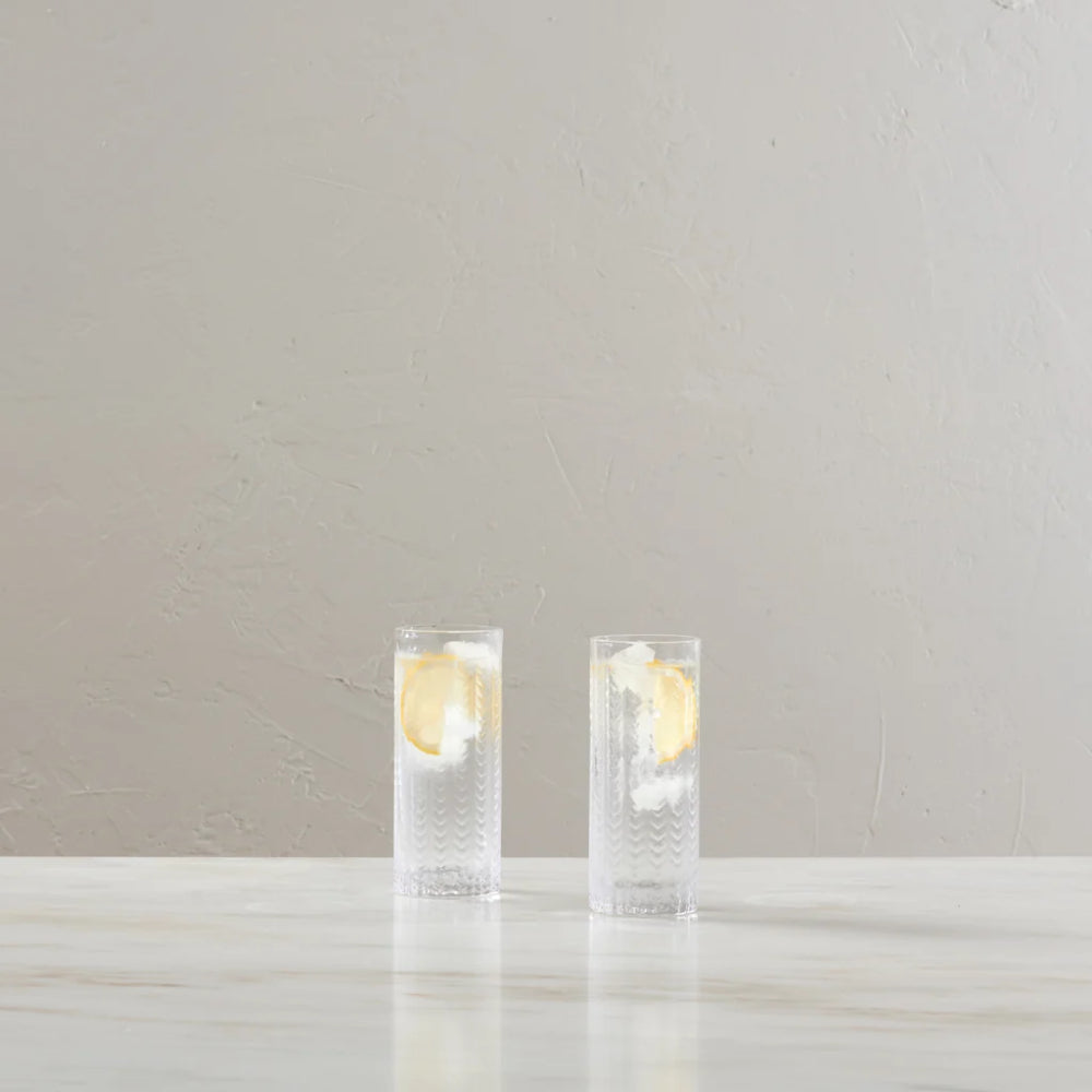 Two clear textured glass tumblers with lemon slices, standing on a light marble surface against a neutral beige wall.