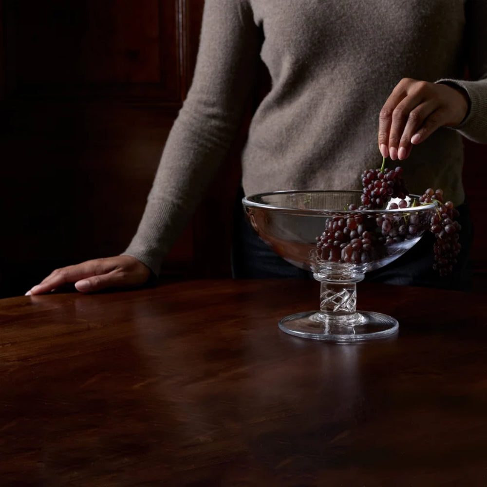 A clear glass pedestal bowl filled with dark red grapes sits on a polished dark wood table, with a person in a taupe sweater reaching in from the side.