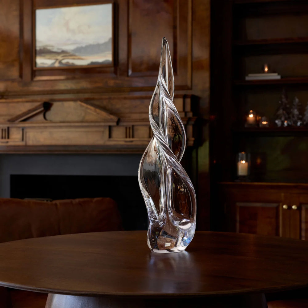 A clear glass sculpture with a twisted, flame-like form stands on a dark wooden table in a warm, dimly lit room with wood paneling and a fireplace in the background.