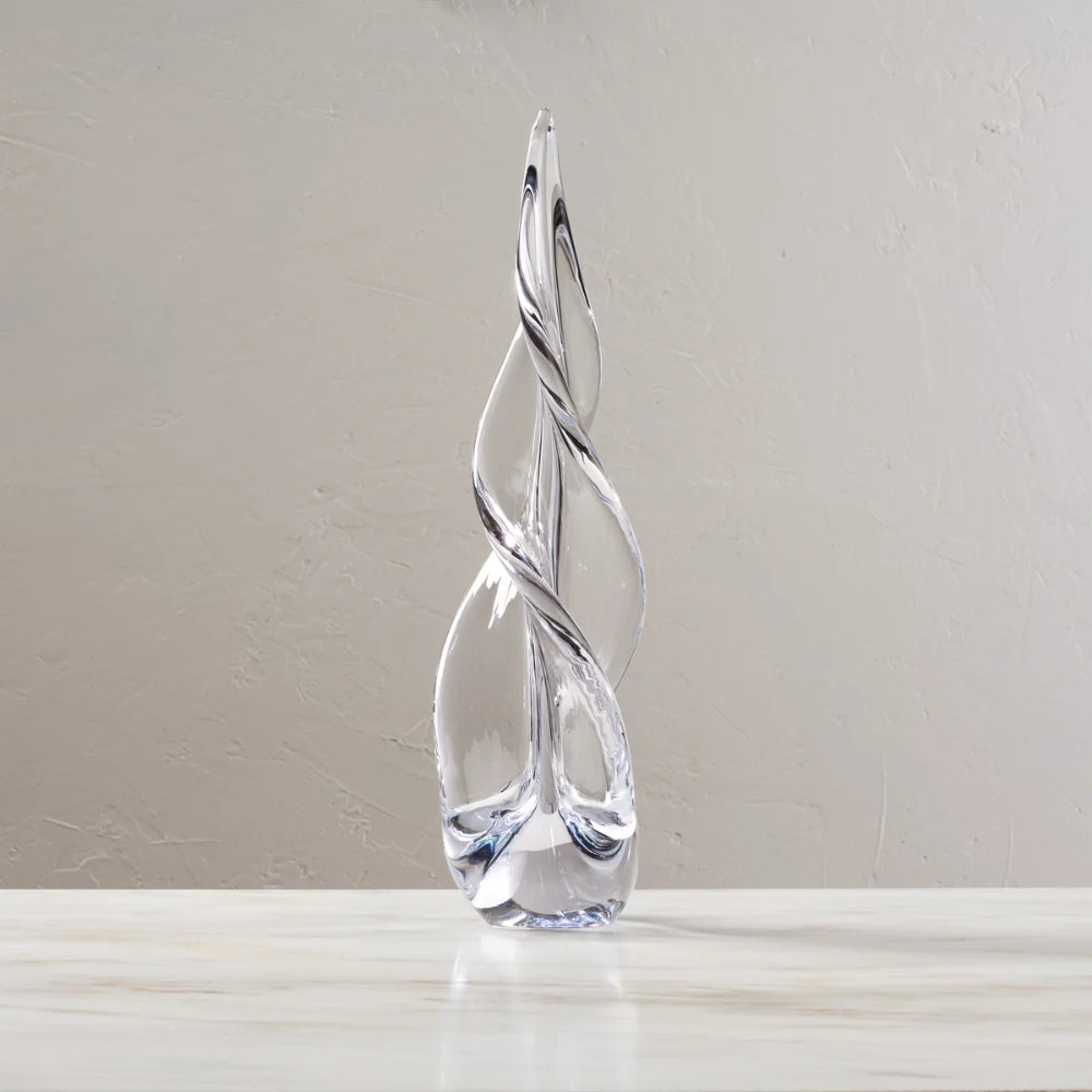 A clear glass sculpture with a twisted, tree-like form standing on a light marble surface against a neutral beige wall.