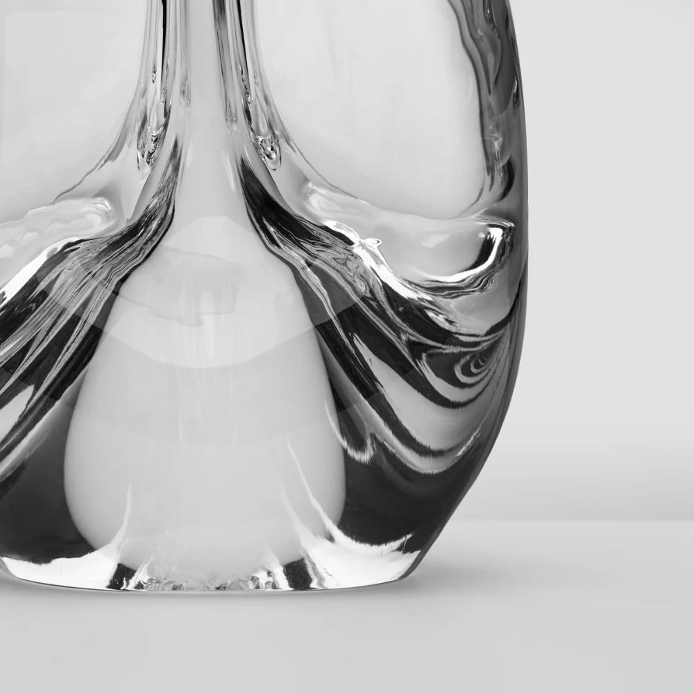 A close-up studio shot of a sculptural clear glass object with flowing, organic wavy forms and reflective highlights on a light gray background.