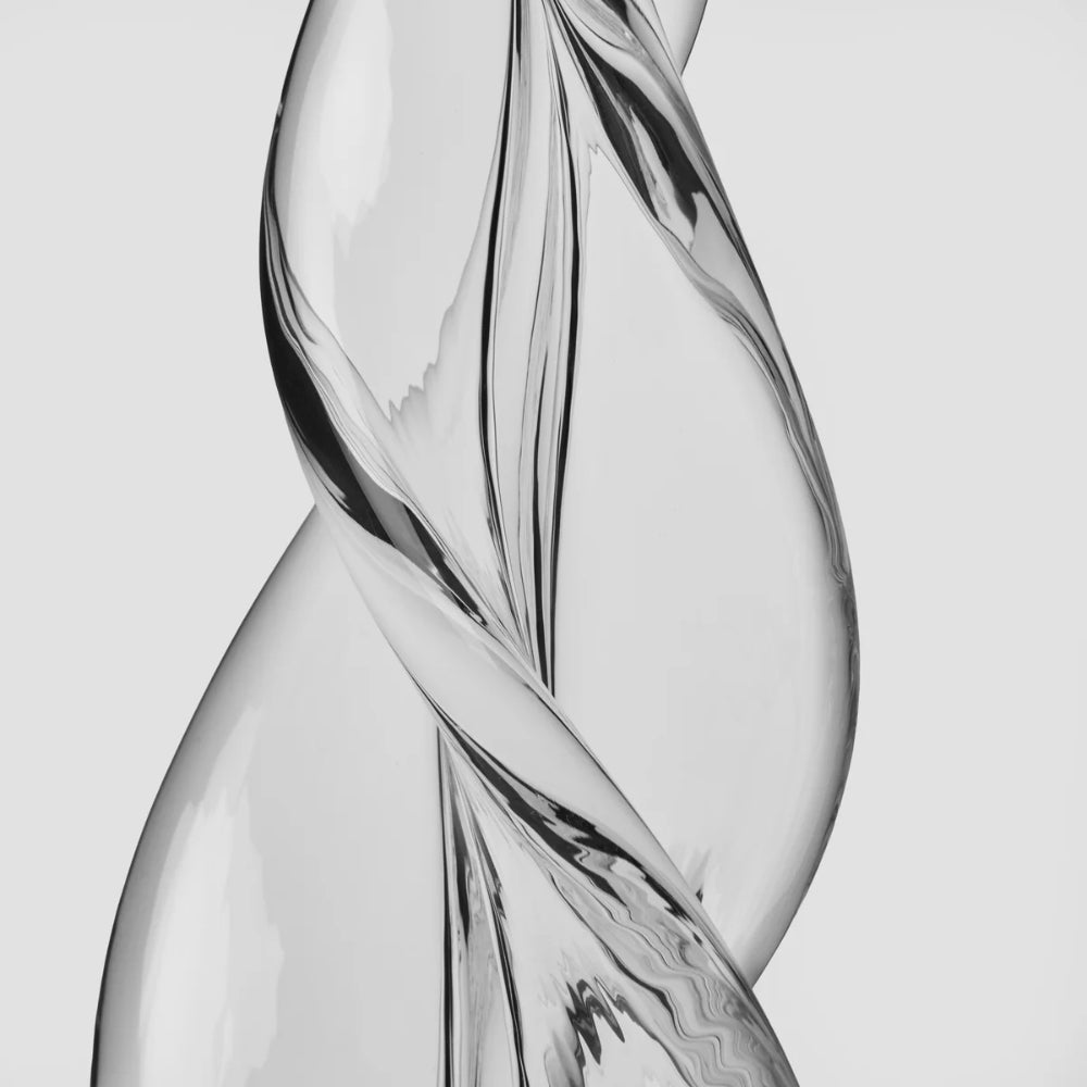 A close-up studio shot of a transparent sculptural glass object with a twisted, flowing form against a light gray background.