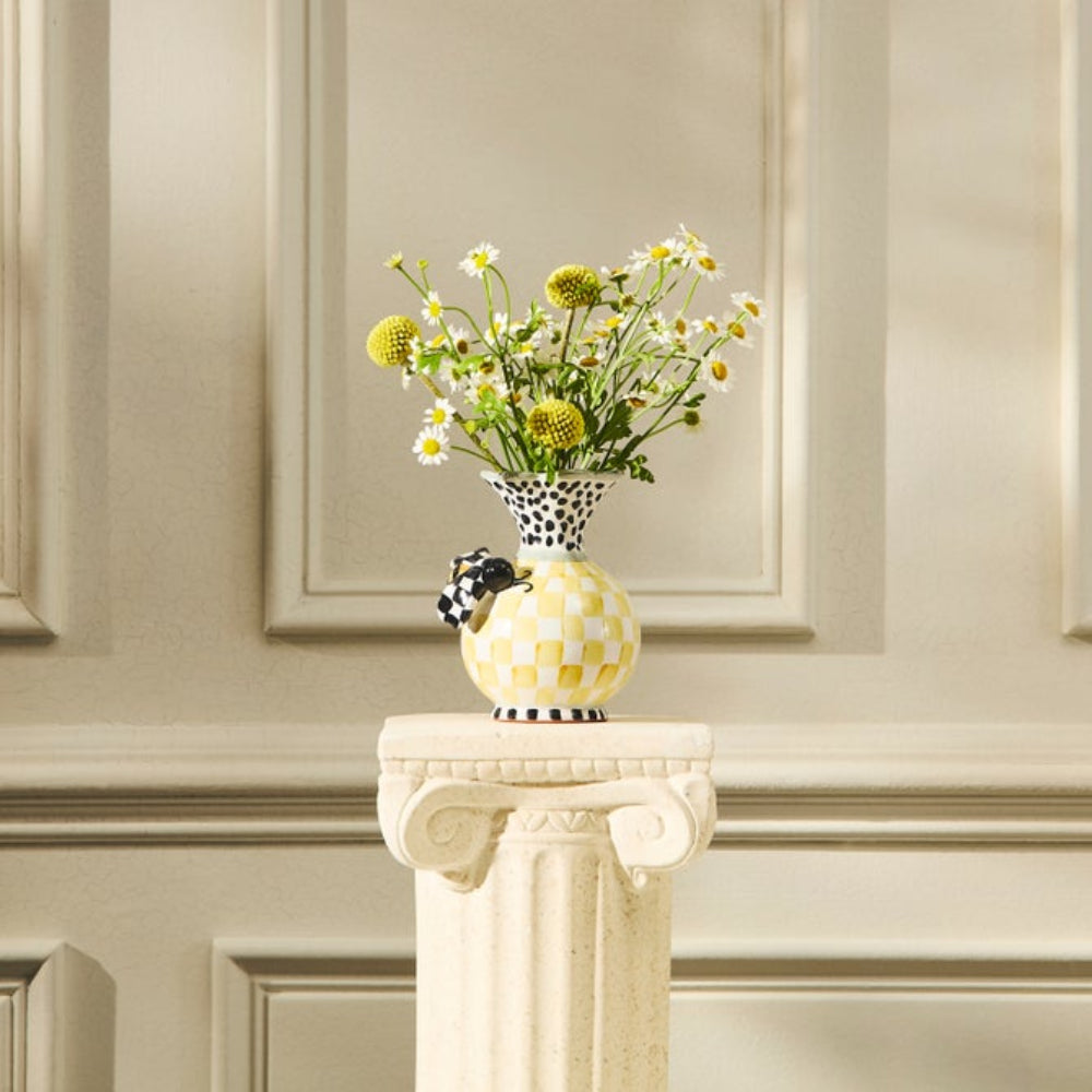  Colorful white, black, green  & yellow bee vase with a bee on the front on a petastal with yellow flowers in it agaist a textered white wall backround. 