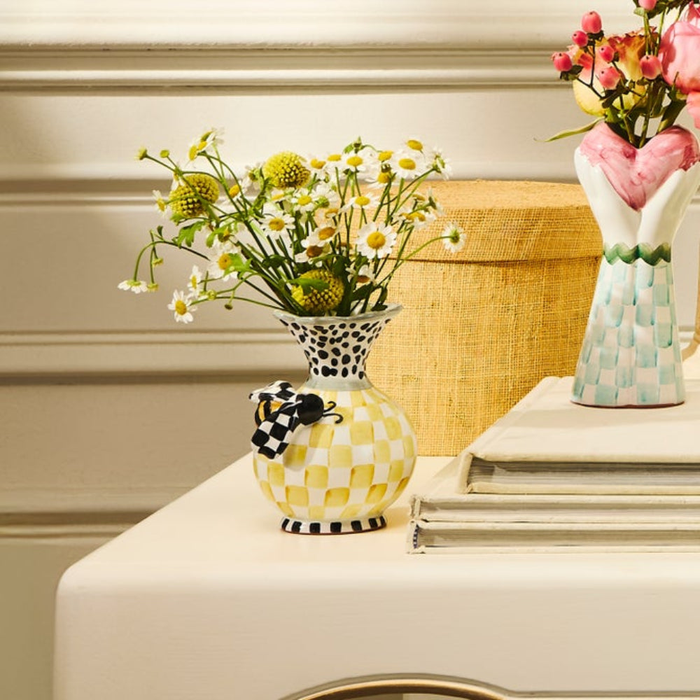 A colorful white, black, green  & yellow bee vase with a bee on the front on a petastal with yellow flowers in it on white tabled clothed table with books and a vase. 