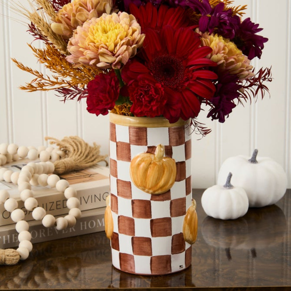 A ceramic vase with a brown and white checkered pattern and small painted pumpkins, filled with a colorful autumn floral arrangement, displayed on a dark wooden table with decorative props.