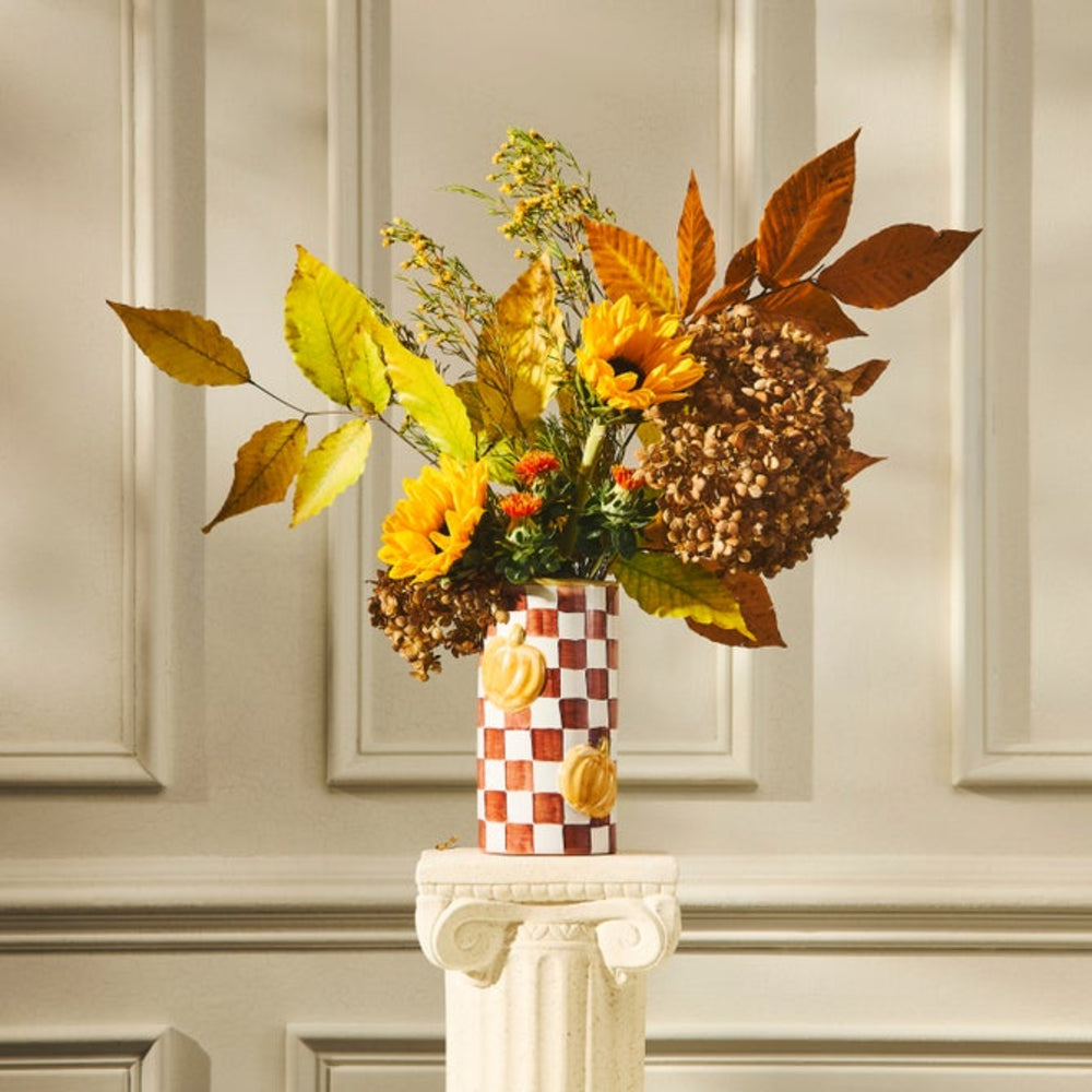 An autumn floral arrangement in a checkered ceramic vase, displayed on a white classical-style pedestal against a beige paneled wall.