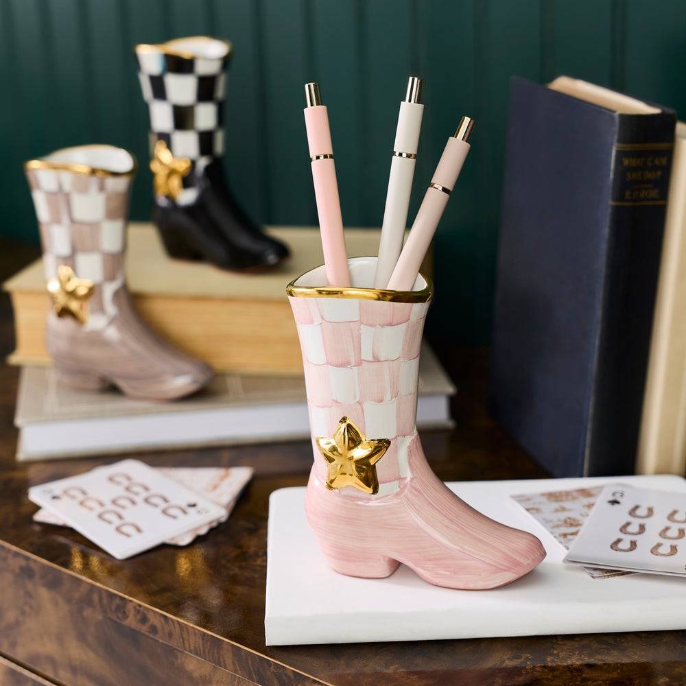 A pink cowboy boot-shaped pen holder with gold trim and a gold star ornament, holding three pastel pens, styled on a wooden table with books and other boot-shaped decor in the background.