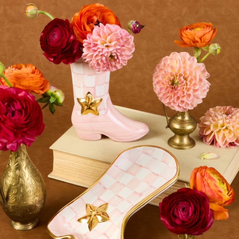 A decorative boot-shaped dish in pale pink with a gold star accent, styled with flowers and placed on a book against a warm brown background.