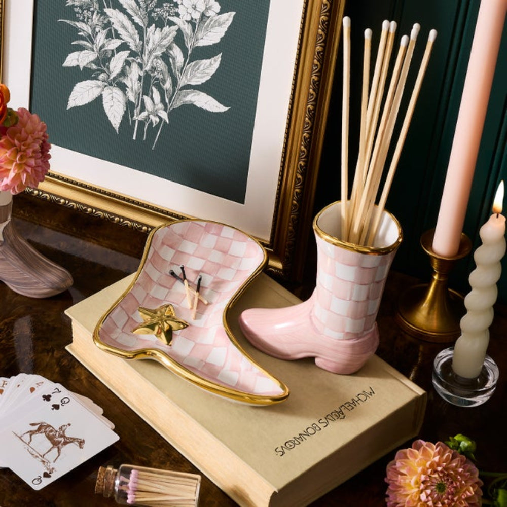 A styled tabletop scene featuring a pink and white checkered boot-shaped incense holder with gold trim, alongside a matching boot-shaped reed diffuser cup holding wooden diffuser sticks. The set is arranged on a book with candles, flowers, cards, and framed botanical art in the background.