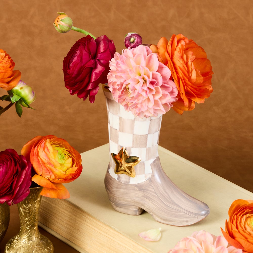 A decorative boot-shaped flower vase with a pink and white checkered upper, a gray footbed, and a gold star ornament, filled with large orange, pink, and burgundy flowers, styled on a book against a brown textured background.