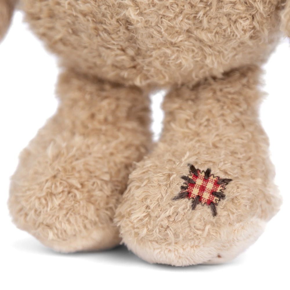 A close up of a beige teddy bear's feet with a red checkared patch on its foot on a white backround. 
