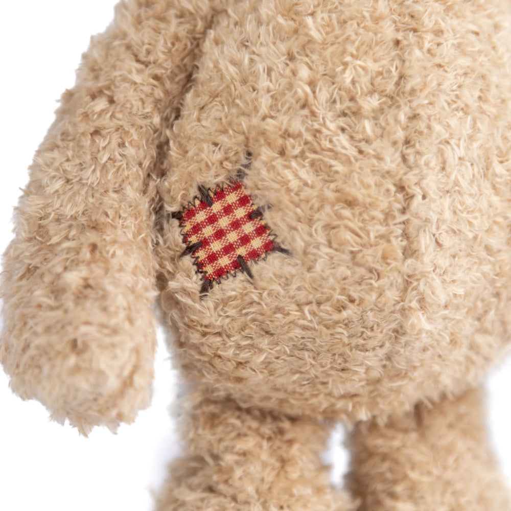 A close up of a beige teddy bear's side with a red checkared patch on its foot on a white backround.