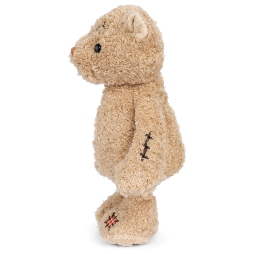 A side photo of a beige stuffed bear standing with a scar on its head and a wound on its side, wearing a red and white checkered shirt, sitting upright with the phrase 'Nobody is Perfect' printed on its foot.