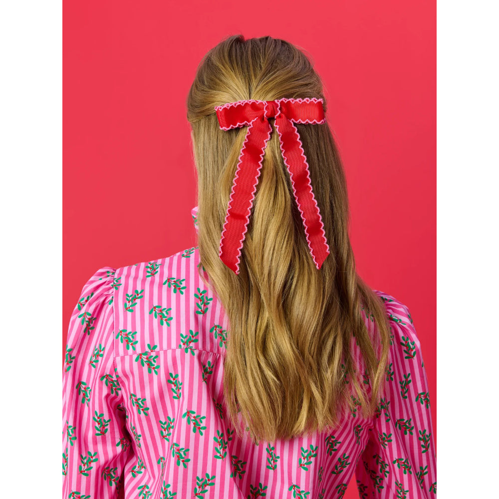 A woman viewed from behind wearing a bright red bow with white scalloped edges in her half-up ponytail. She is wearing a pink and white striped shirt with green floral patterns, set against a solid red background.