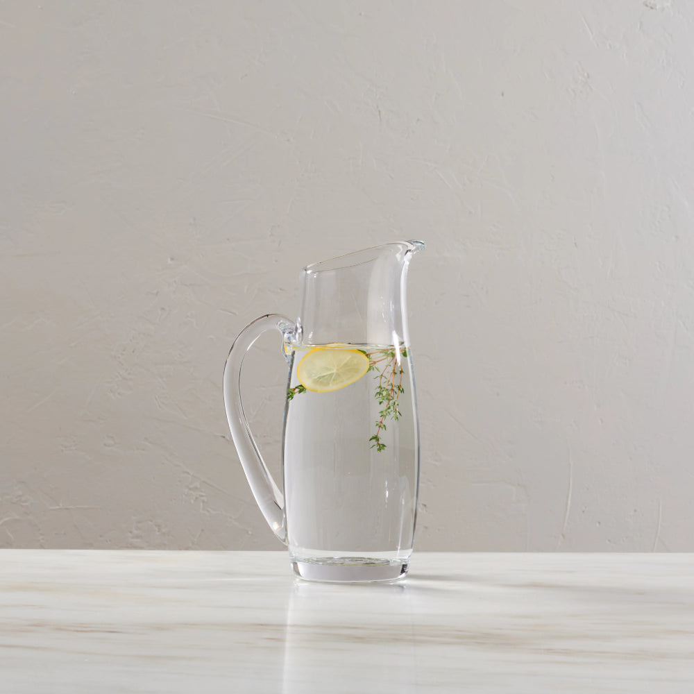 A clear glass pitcher with a handle and spout, filled with water and containing a lemon slice and herbs, set on a light marble surface against a neutral textured wall.