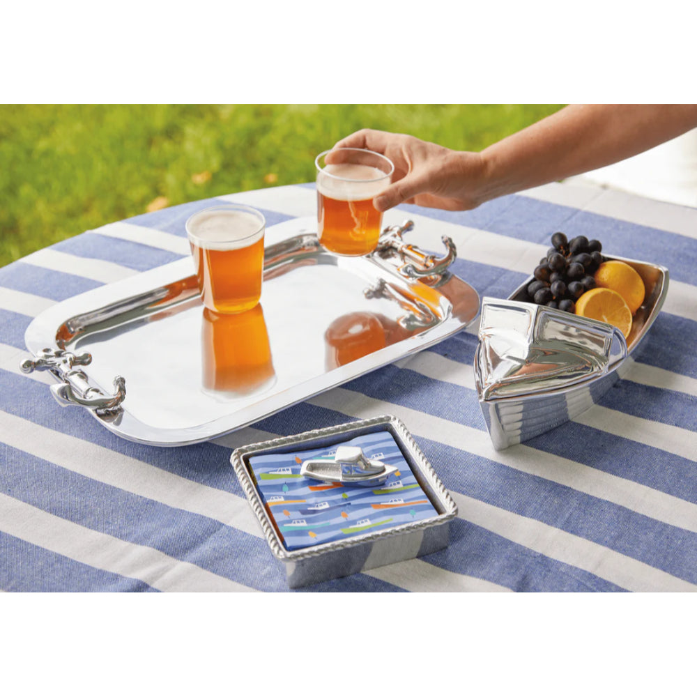 Silver tray with silver anchor handles with drinks on it, on  a blue and white striped table clothed table with a silver boat and silver boxed napkin holder with blue napkins outside. 