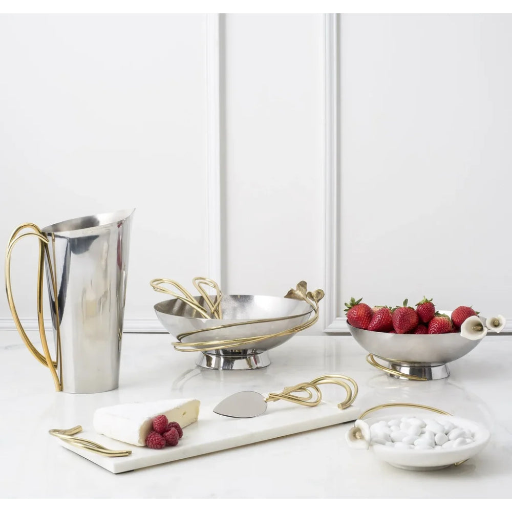 A coordinated set of metallic serving pieces on a white surface: a tall silver pitcher with a gold handle, two silver bowls with gold trim and handles, a white serving board with a silver cheese knife, strawberries, and a small dish of white candies. The background is a clean white interior wall with panel lines.