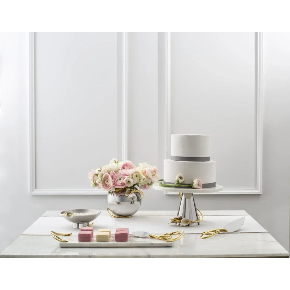 A styled tabletop scene with a two-tier white cake on a silver pedestal stand, a vase of pink and white flowers, small pink and cream cubes on a tray, and gold ribbon accents against a white paneled wall background.