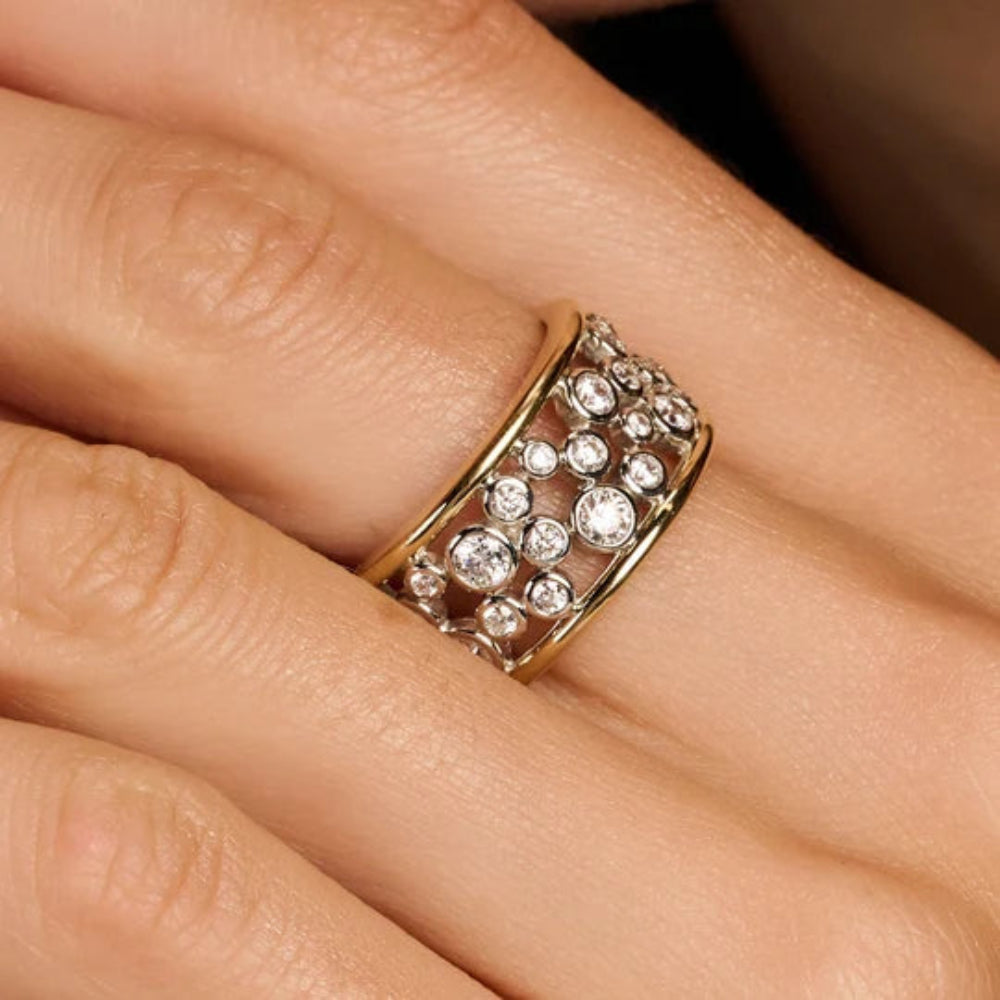 A close-up of a gold band ring adorned with multiple rows of clear round gemstones, worn on a person's finger.