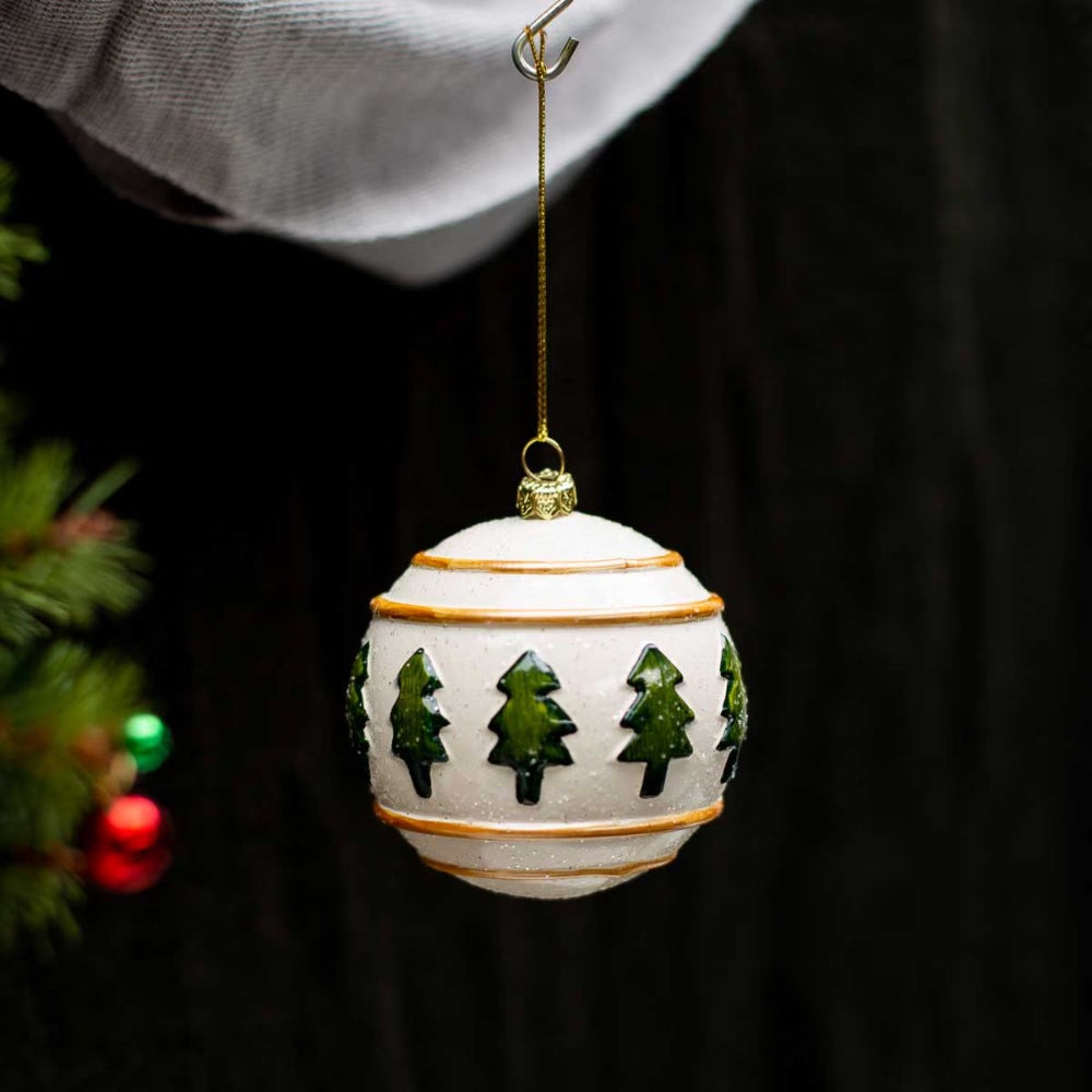 A white ceramic Christmas ornament hanging from a gold string, decorated with green tree silhouettes and thin gold bands, set against a dark background with blurred greenery and lights on the left.