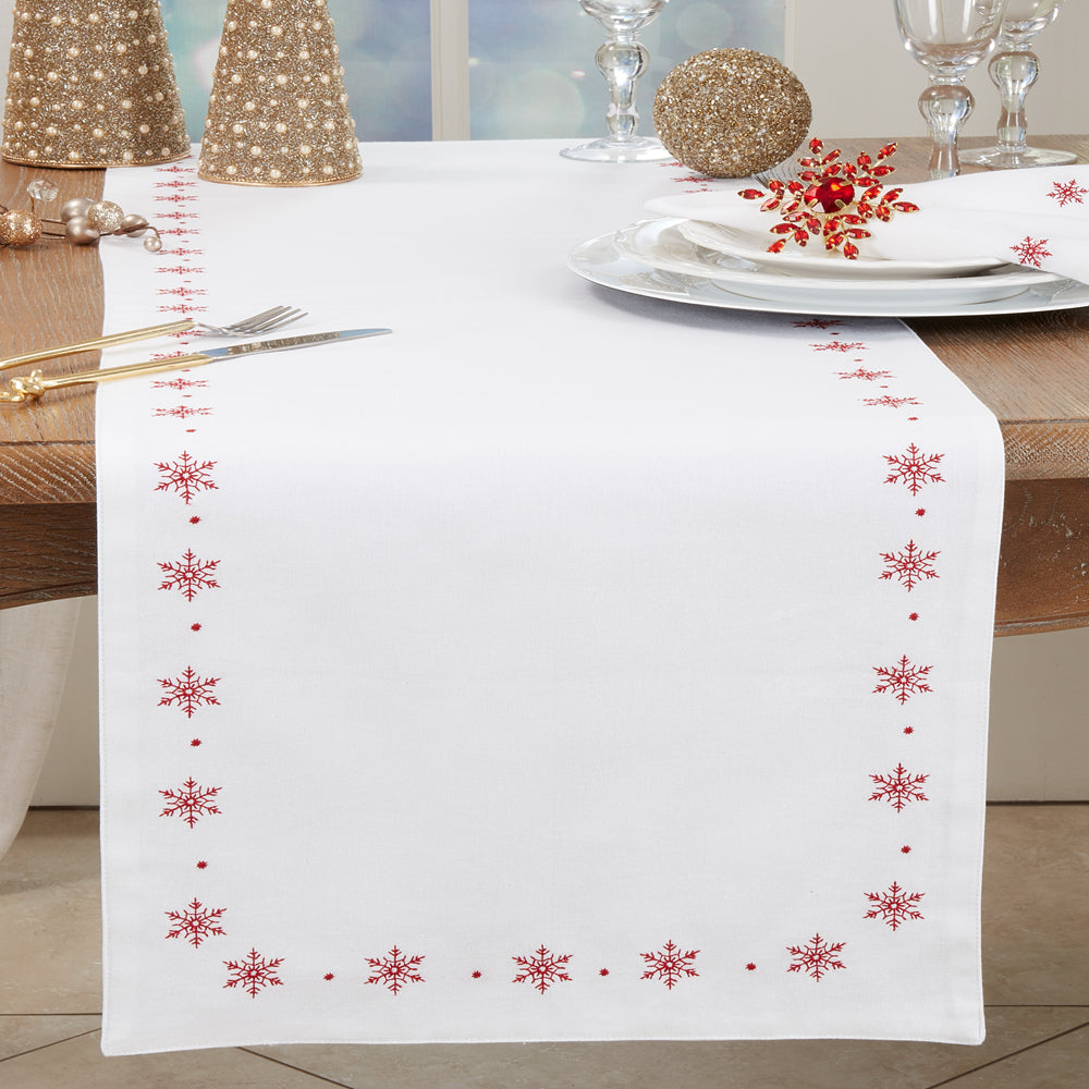 A white table runner with a border of red embroidered snowflakes and dots, styled on a wooden dining table with plates, cutlery, and holiday decorations.