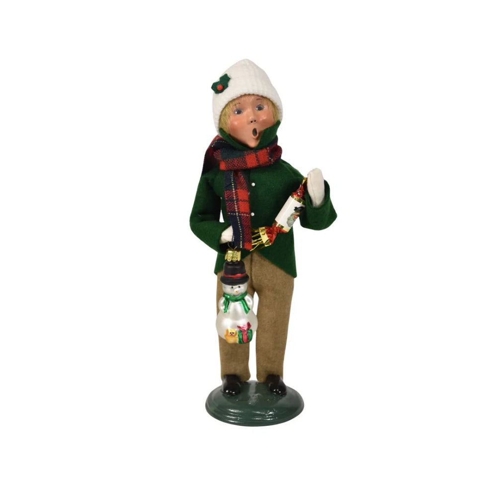 Christmas figurine of a boy caroling in winter clothing in a green jacket, tan paints, red plaid scarf and white cap holding a with a snowman ornament and popper on a white background