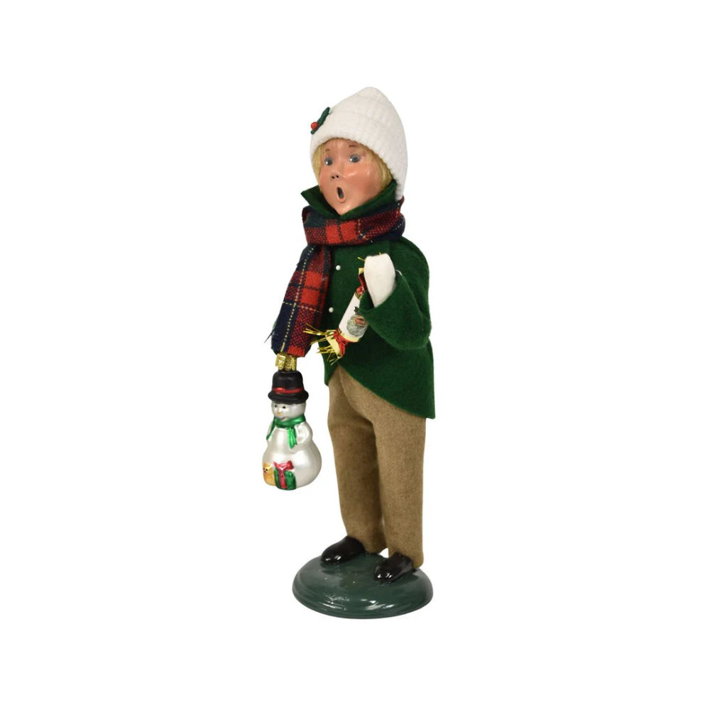 An left side angled photo of a christmas figurine of a boy caroling in winter clothing in a green jacket, tan paints, red plaid scarf and white cap holding a with a snowman ornament and popper on a white background
