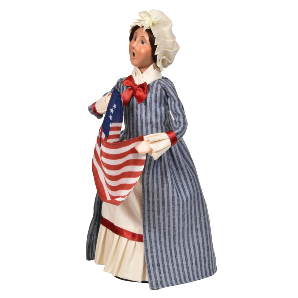 Left side photo of a figurine of a caroling Betsy Ross in historical attire holding an American flag on a white background