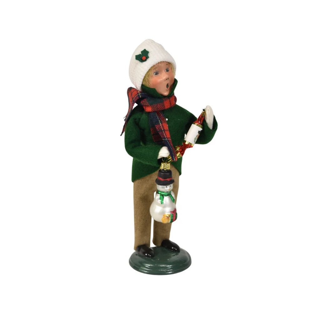 An right side angled photo of a christmas figurine of a boy caroling in winter clothing in a green jacket, tan paints, red plaid scarf and white cap holding a with a snowman ornament and popper on a white background