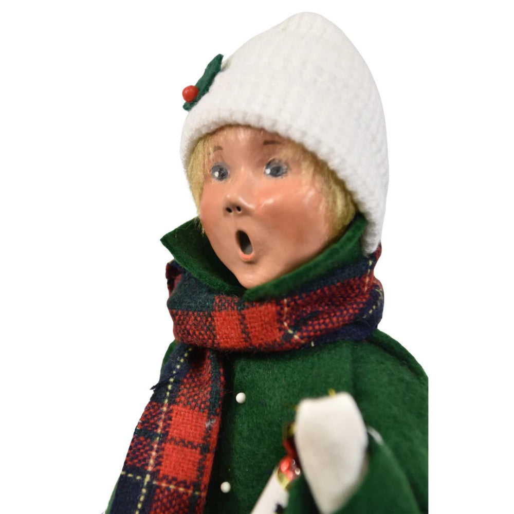 A close up photo of the face of a  christmas figurine of a boy caroling in winter clothing in a green jacket, tan paints, red plaid scarf and white cap holding a with a snowman ornament and popper on a white background