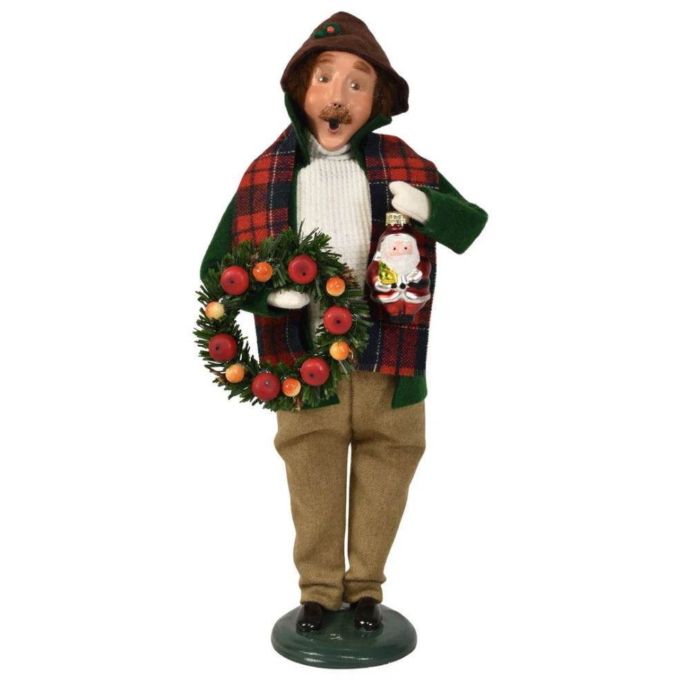 A figurine of a caroling man holding a christmas wreath and a small santa ornament in a vintage-style red plaid scarf and green coat and white knitted sweater and tan pants on a white background