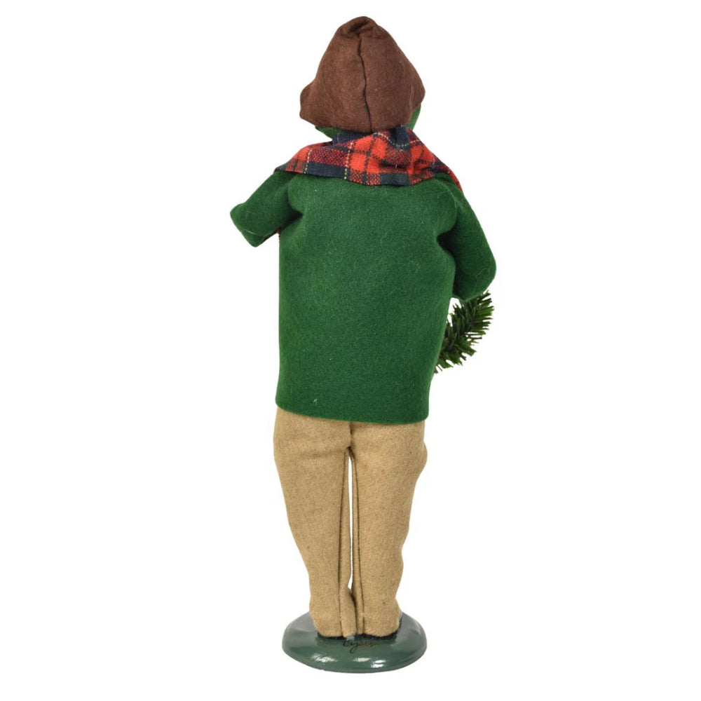 A  photo of the back  a decorative figurine of a caroling man holding a christmas wreath and a small santa ornament in a vintage-style red plaid scarf and green  coat and white knitted sweater and tan pants on a white background