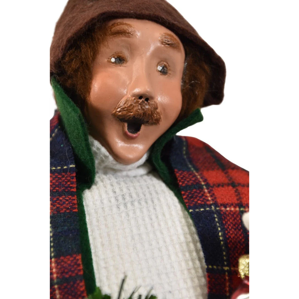 A close photo the face of a decorative figurine of a caroling man holding a christmas wreath and a small santa ornament in a vintage-style  red plaid scraf green jacket  and white knitted sweater on a white background