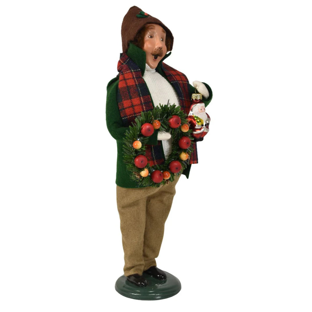 An right angled photo of a decorative figurine of a caroling man holding a christmas wreath and a small santa ornament in a vintage-style red plaid scarf and green  coat and white knitted sweater and tan pants on a white background