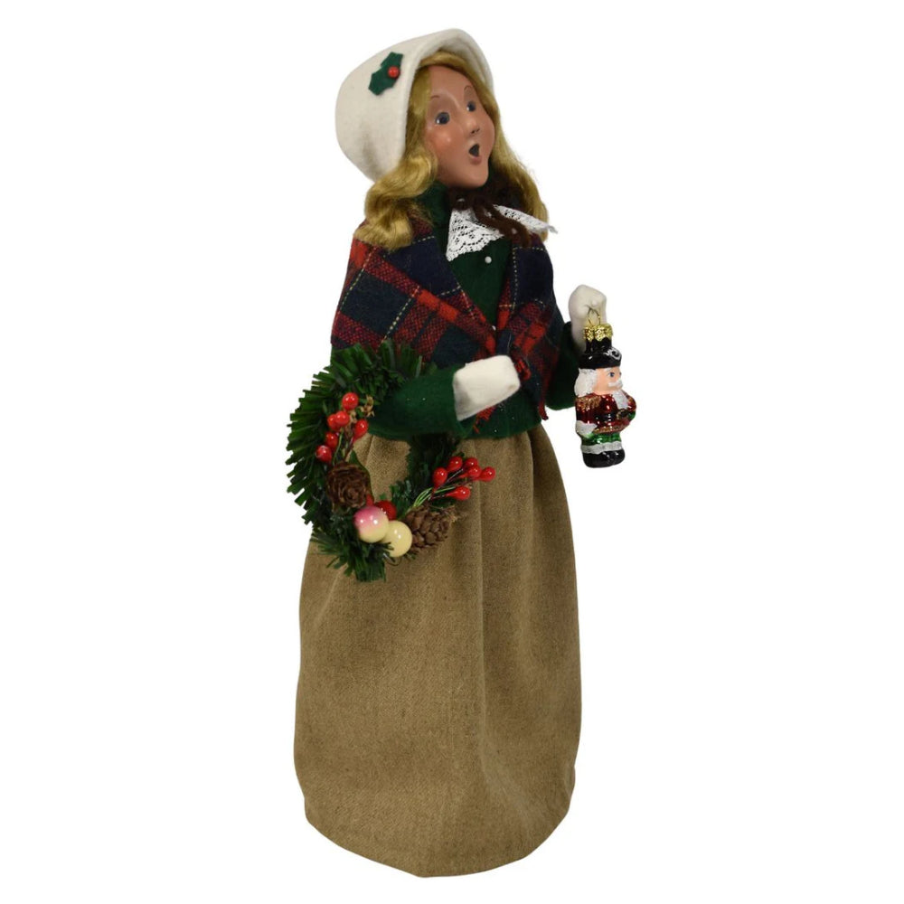 Side angled photo of a colorful holiday decorative figurine of a  caroling woman holding a wreath and a small nutcracker ornanment  on a white background