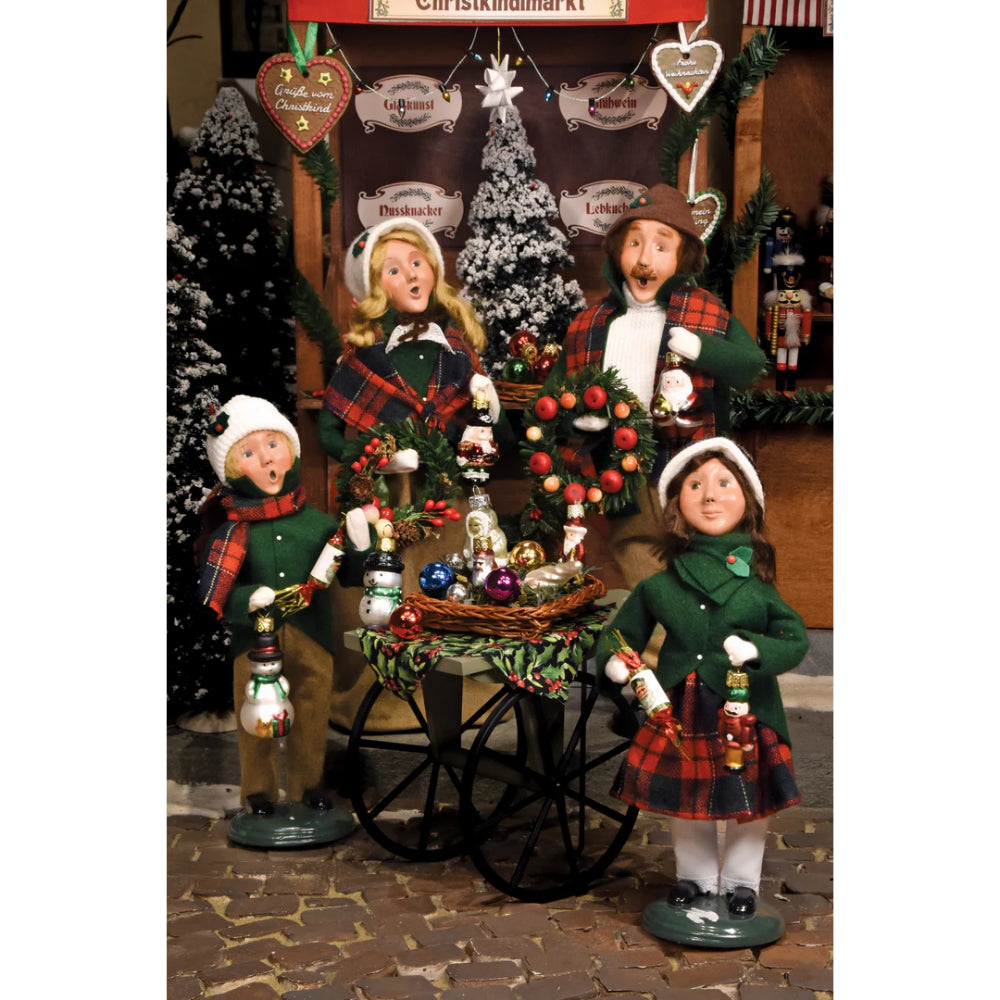 Christmas-themed family figurines with a cart full of ornaments and trees in a festive setting.