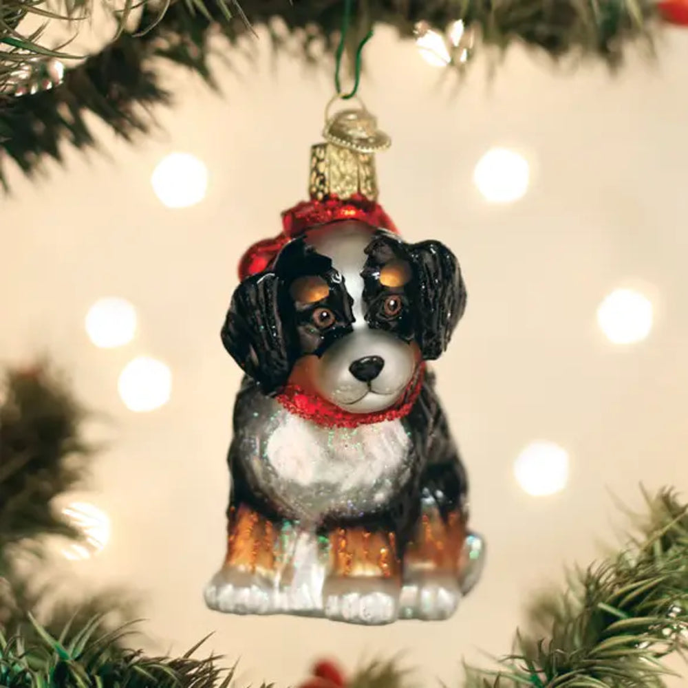 A Bernese mountain dog Christmas ornament hanging from a green tree branch, with warm white bokeh lights in the background.