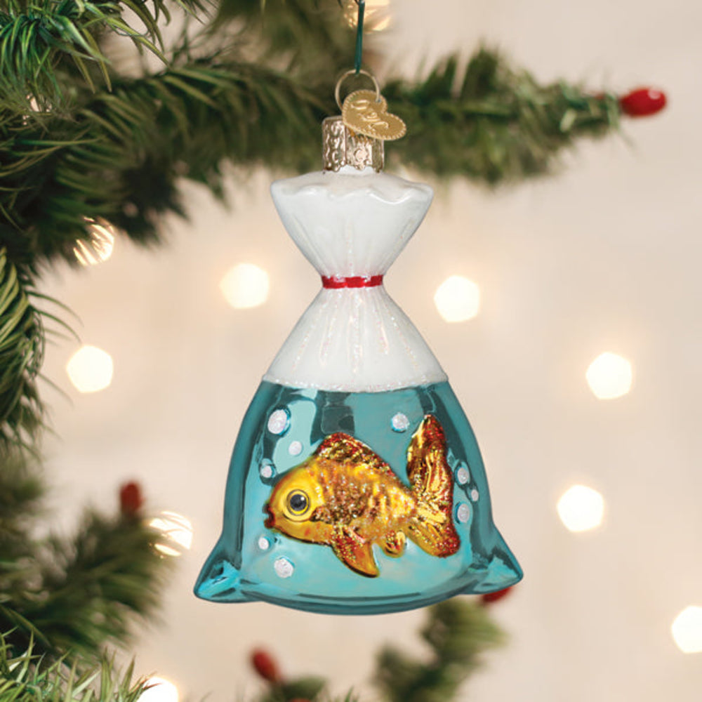 A Christmas ornament shaped like a glass bag filled with blue water and a goldfish, hanging from a green Christmas tree branch with warm blurred lights in the background.