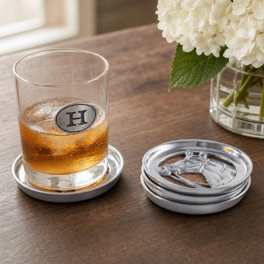 A clear rocks glass with a silver monogrammed 'H' coaster sits on a coaster next to stacked silver coaster lids on a wooden table, with white flowers in the background.
