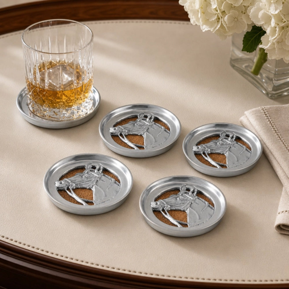 Four round silver metal coasters with an embossed horse design, arranged on a beige tablecloth alongside a glass of whiskey and white flowers.