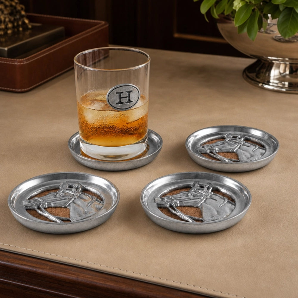 Four silver-toned metal whiskey coasters with embossed horse designs arranged on a tan leather tabletop, with one coaster holding a glass of amber liquor and ice. A monogrammed glass is visible on the coaster.