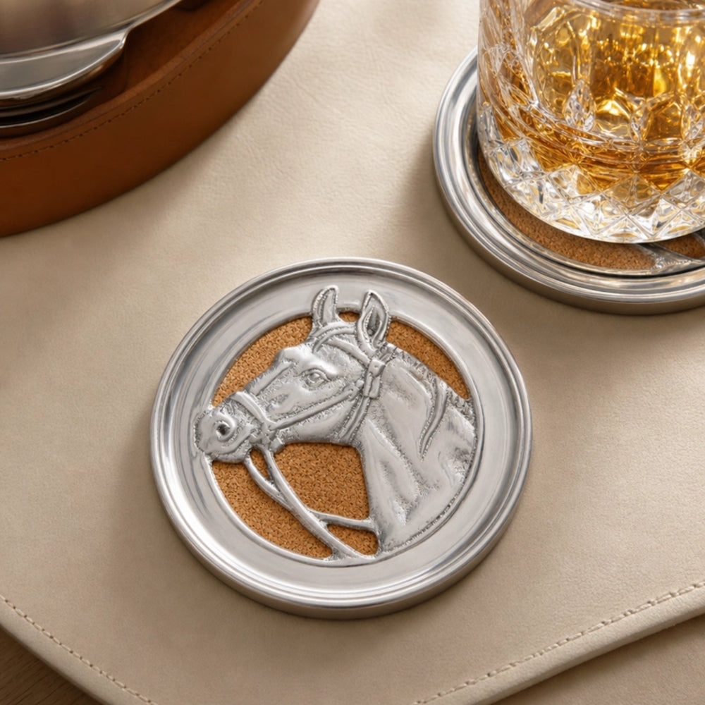 A round silver-toned coaster featuring an embossed horse head design over a tan cork-like center, shown on a beige leather surface next to a glass of amber liquid on a matching coaster.