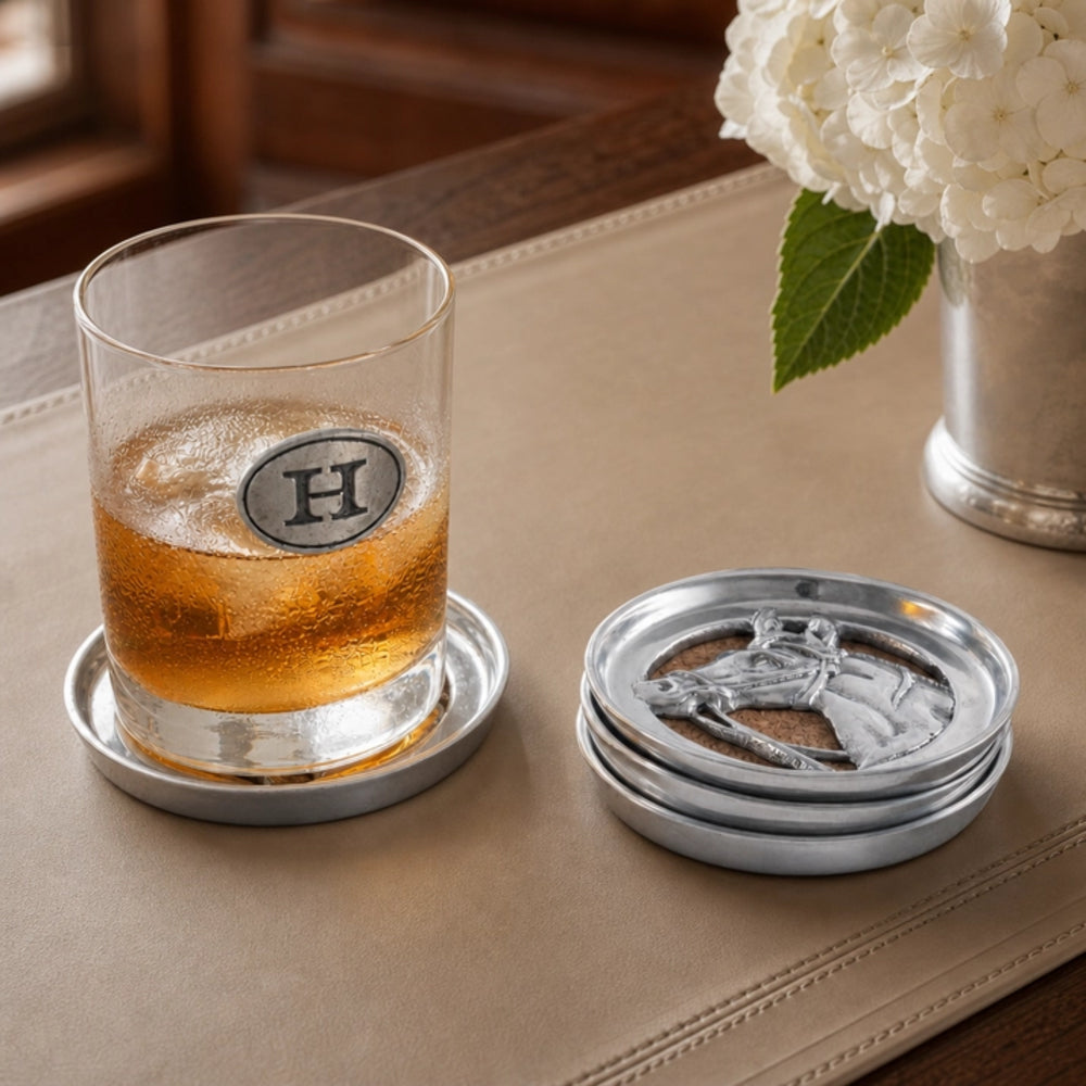 A clear rocks glass with a silver monogrammed 'H' coaster sits on a silver coaster, next to a stack of three matching coasters. The set is styled on a beige leather tray with a glass of amber whiskey and white flowers in the background.