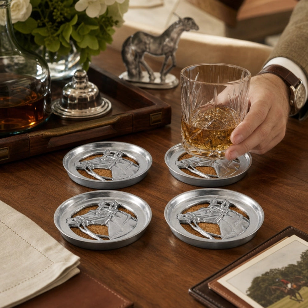 Four silver horse-head coasters on a wooden table, with a hand holding a glass of whiskey over one coaster. A bottle, flowers, and a decorative horse figurine are visible in the background.