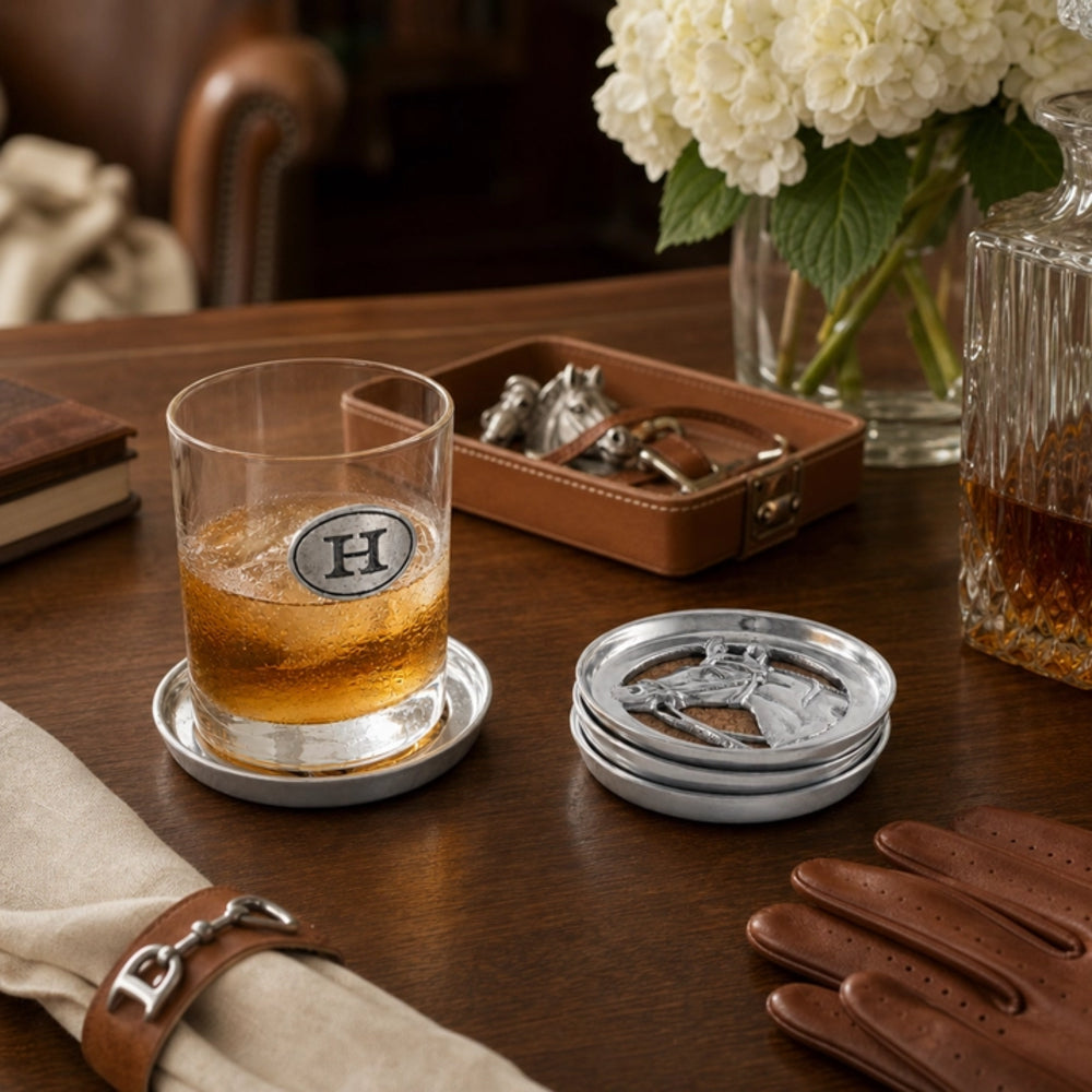 A clear glass whiskey tumbler with a circular silver coaster engraved with a monogram letter H, filled with amber liquor, sits on a silver coaster on a dark wood table. A stack of matching silver coasters is beside it, with leather gloves, a rolled napkin, a brown leather case, and a glass vase of white flowers in the background.