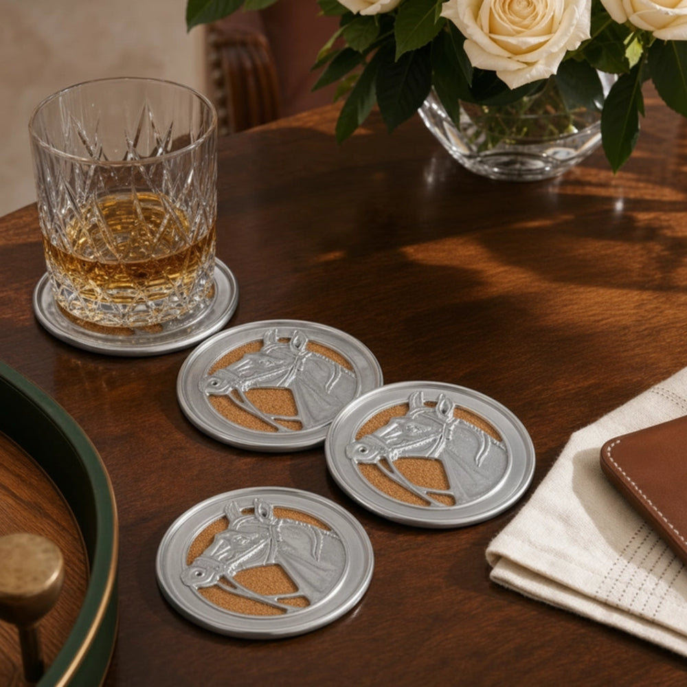 Four round silver horse-head coasters with tan cork inserts are arranged on a dark wooden table, alongside a whiskey glass on a coaster and a vase of white roses in the background.