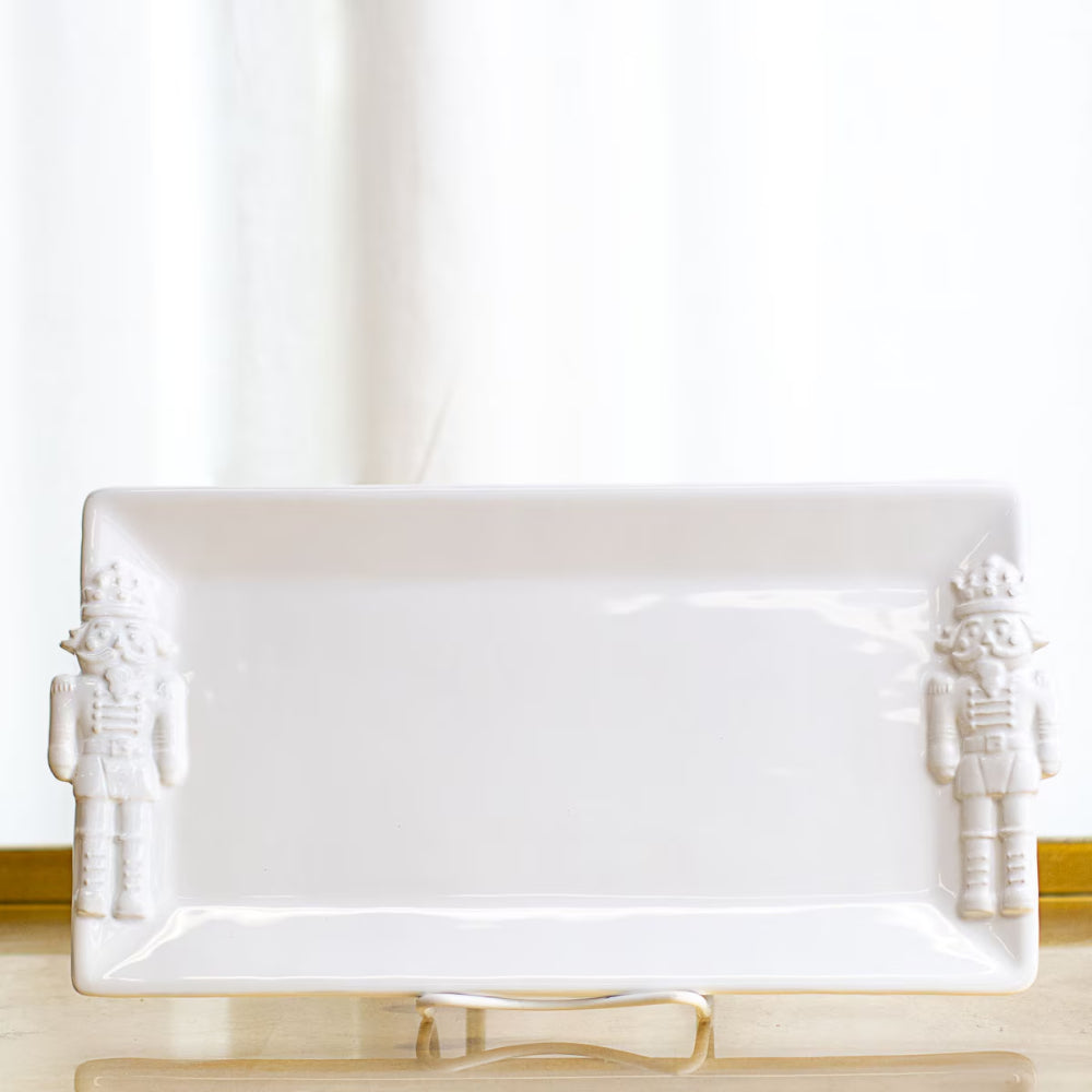 A white rectangular ceramic serving platter with embossed nutcracker figures on both short ends, displayed on a stand against a soft white curtain background.