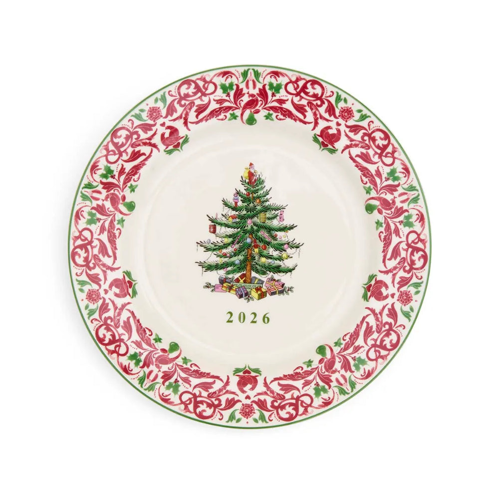 A round ceramic Christmas plate with a white center, featuring a green Christmas tree and presents illustration in the middle and a red floral scroll border with green accents around the rim. The year '2026' is printed below the tree.
